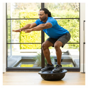 Strength Training Equipment- (300*300)- Man with gym outfit performing a squat using a Black Bosu Ball (inverted) in an indoor setting with panoramic windows with a small pond and tall hedge visible outside