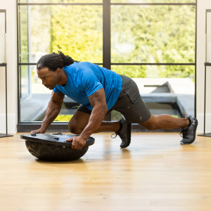Strength Training Equipment- (300*300)- Man with gym outfit performing mountain climbers using a Black Bosu Ball (inverted) in an indoor setting with panoramic windows with a small pond and tall hedge visible outside
