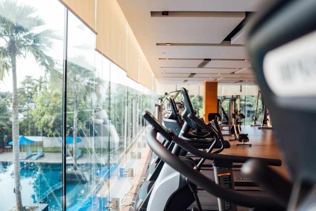 A spacious modern gym with a row of treadmills and other exercise equipment facing large windows overlooking a lush outdoor area with a swimming pool and palm trees, ideal for a motivating workout experience in Auckland, New Zealand.